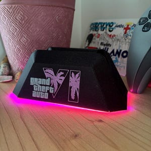 May include: A black, trapezoidal gaming controller stand featuring the Grand Theft Auto VI logo and palm tree graphics in a light purple color. The base of the stand emits a bright, vibrant pink LED glow that illuminates the surface below it.