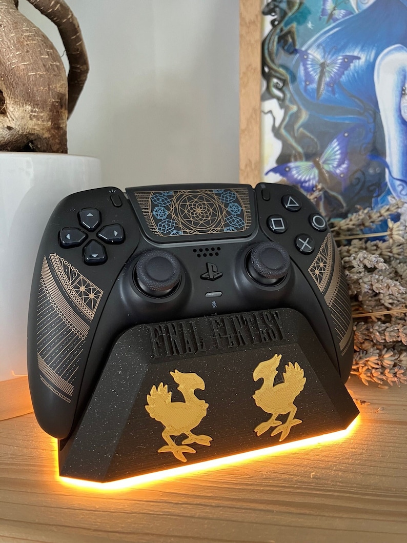 K&ouml;nnte beinhalten: Ein schwarzer PlayStation 5 DualSense-Controller mit einem goldenen und blauen geometrischen Design auf dem Touchpad. Der Controller sitzt auf einem schwarzen Ladest&auml;nder mit einem goldenen Final Fantasy-Logo und zwei goldenen Chocobo-Figuren. Der St&auml;nder hat ein leuchtend orangefarbenes Licht um die Basis.