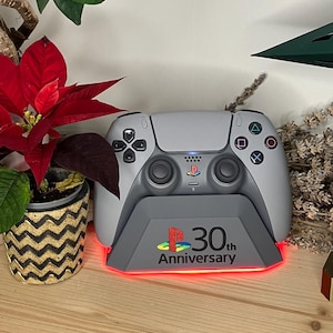Op de afbeelding: Een grijze PlayStation 5-controller op een oplaadstation met een rode, oplichtende basis. Het oplaadstation heeft het PlayStation-logo en de tekst "30th Anniversary". Decoratieve planten omringen de controller.