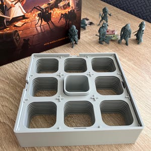 Op de afbeelding: Een grijze plastic spelorganizer met meerdere vierkante compartimenten en een centrale rechthoekige ruimte. Miniatuur spelfiguren liggen verspreid rond de organizer, met een speldoos met fantasiekunst op de achtergrond.