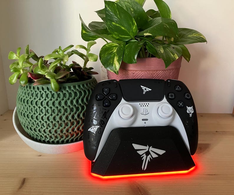 May include: A black and white video game controller with a charging stand. The stand has a red light around the base and a white firefly logo. The controller has a white firefly logo and a wolf logo.