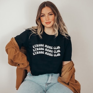 May include: Black t-shirt with the text "Strong Moms Club" repeated three times in white, wavy letters. The shirt is worn by a person with long brown hair and is paired with blue jeans and a brown jacket.