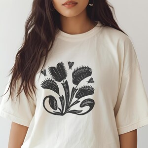 Può includere: T-shirt color crema con grafica nera di una pianta carnivora Venere acchiappamosche con diverse trappole aperte e mosche stilizzate. Il disegno è centrato sul davanti della maglietta. La maglietta ha maniche corte e girocollo.