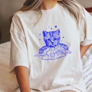 Op de afbeelding: Crèmekleurig T-shirt met een blauwe illustratie van een katje op een wolk en sterren. De afbeelding staat in het midden van de voorkant. Het shirt heeft korte mouwen en een relaxte pasvorm.