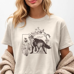 Puede incluir: Camiseta beige con una ilustración detallada de un zorro dentro de un marco rectangular. La obra de arte incluye mariposas, flores y ramas, creando un diseño inspirado en la naturaleza. La camiseta tiene mangas cortas y cuello redondo.