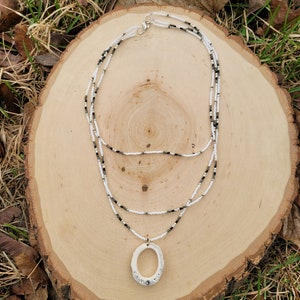 May include: A layered necklace with white and black beads and a large, oval pendant with rhinestones. The necklace is displayed on a wooden surface.