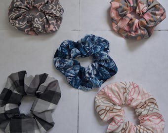 May include: Five fabric scrunchies in various patterns and colors. One is gray with a floral design, another is pink and gray with a floral pattern, and a third is blue with a floral pattern. The other two are checkered and geometric.