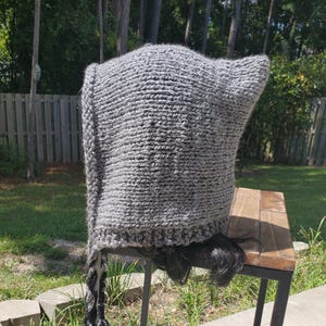 May include: A gray knitted hooded hat with a long, braided cord. The hat is sitting on a wooden table.