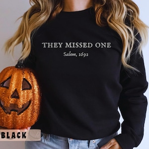 Puede incluir: Sudadera negra con el texto "They Missed One Salem, 1692" en letras blancas.