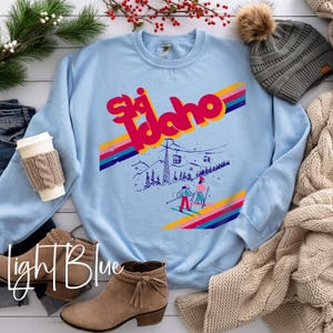 May include: Light blue sweatshirt with the text "Ski Idaho" in a retro font, featuring a ski lift and skiers. The sweatshirt is surrounded by a gray beanie with a pom-pom, a beige cable-knit blanket, and brown boots.