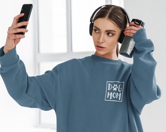 May include: A woman wearing a blue sweatshirt with the text "DOG MOM" and a paw print in a white box. She is holding a phone in one hand and a water bottle in the other.