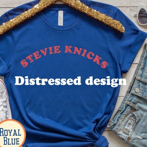Puede incluir: Una camiseta de manga corta color azul real con el texto STEVIE KNICKS en una fuente roja desgastada arqueada sobre el pecho. La prenda tiene cuello redondo y corte est&aacute;ndar, presentada sobre una percha con lentejuelas doradas.