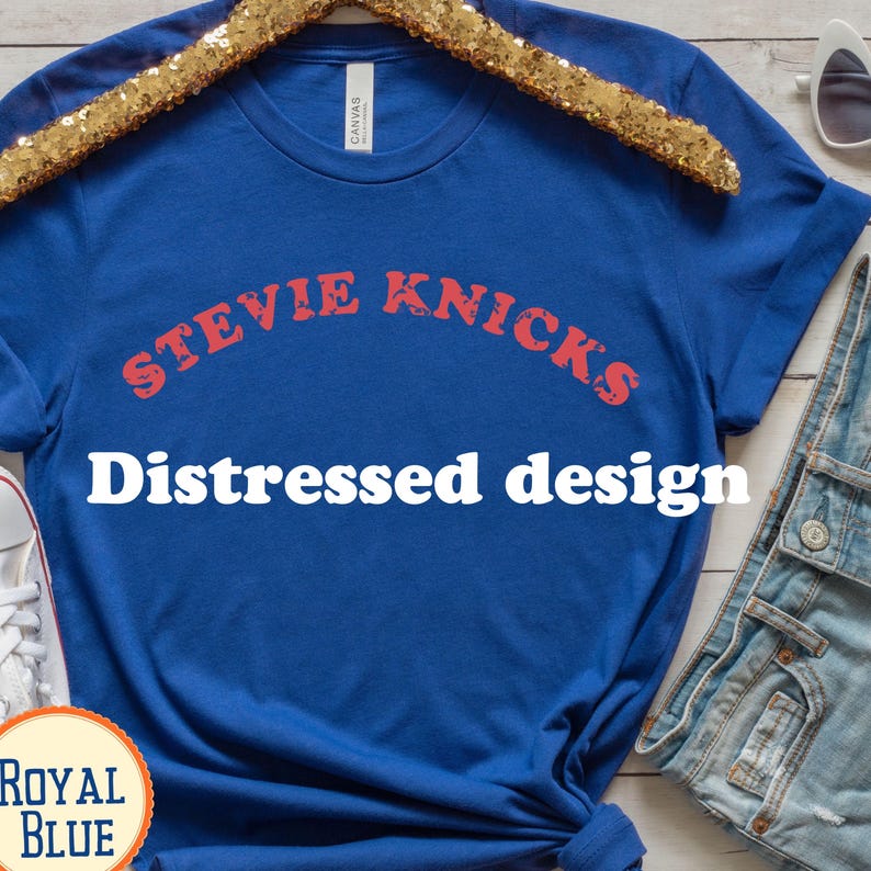Puede incluir: Una camiseta de manga corta color azul real con el texto STEVIE KNICKS en una fuente roja desgastada arqueada sobre el pecho. La prenda tiene cuello redondo y corte est&aacute;ndar, presentada sobre una percha con lentejuelas doradas.