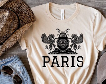 May include: A beige t-shirt with a black graphic of a lion's head with wings on either side. The text "PARIS" is printed below the graphic.