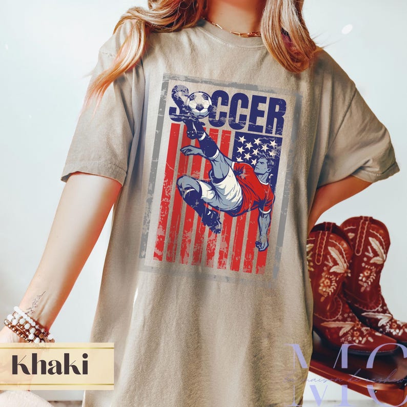 May include: Khaki-colored t-shirt featuring a distressed graphic of a soccer player kicking a ball, set against a backdrop of the American flag. The word "SOCCER" is printed above the player. The shirt is paired with brown cowboy boots.