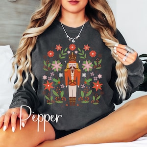 May include: A dark gray sweatshirt with a festive design featuring a nutcracker surrounded by flowers and greenery. The text "Pepper" is visible at the bottom of the image.