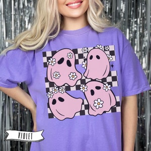 May include: Violet-colored t-shirt featuring a graphic of four pink ghosts adorned with daisy crowns, set against a black and white checkered background. The word "VIOLET" is displayed in a banner at the bottom.