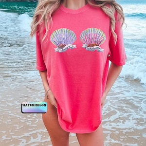 May include: A coral-colored t-shirt featuring two seashell graphics with the text "Lacey's Bachelorette". The seashells are detailed with shades of purple, blue, and yellow. The word "WATERMELON" is printed on a small rectangle at the bottom.