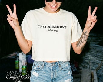 May include: An ivory short-sleeved crop top t-shirt featuring the black text "THEY MISSED ONE" and "Salem, 1692" printed on the chest. The shirt is paired with light blue denim shorts that have a distressed, frayed hem and ripped details.