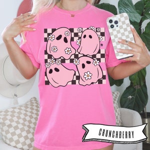 May include: A bright pink t-shirt featuring a graphic of four pink ghosts with daisy crowns on a black and white checkered background. The shirt has the word "CRUNCHBERRY" in a banner at the bottom.