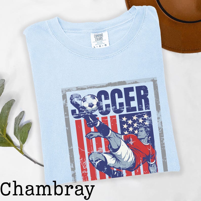 May include: Light blue t-shirt with a graphic of a soccer player kicking a ball, set against a distressed American flag design. The word "SOCCER" is printed above the player. The shirt is labeled "Chambray".