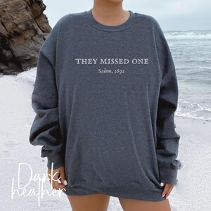 May include: Dark heather grey sweatshirt with the text "They Missed One Salem, 1692" printed on the front.