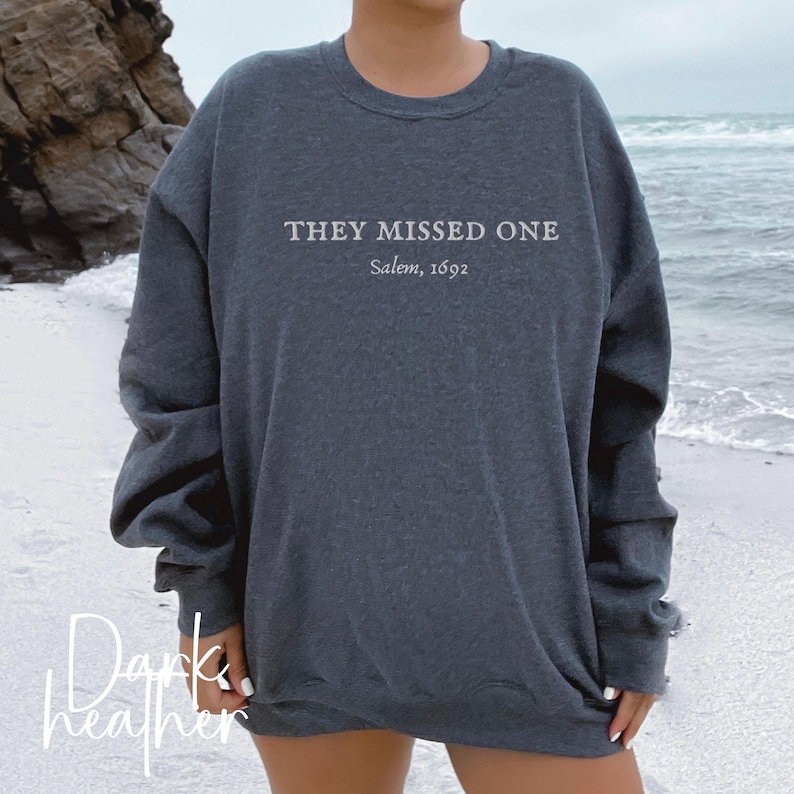 May include: Dark heather grey sweatshirt with the text "They Missed One Salem, 1692" printed on the front.