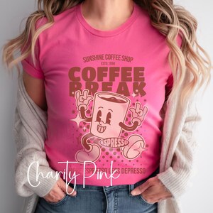 May include: A pink t-shirt with a retro-style illustration of a coffee cup with the text "Sunshine Coffee Shop Est. 1958 Coffee Break Espresso" printed on it.