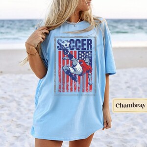 May include: Light blue t-shirt featuring a soccer player kicking a ball, set against a distressed American flag design. The word "SOCCER" is printed above the player. The shirt is a light blue color.