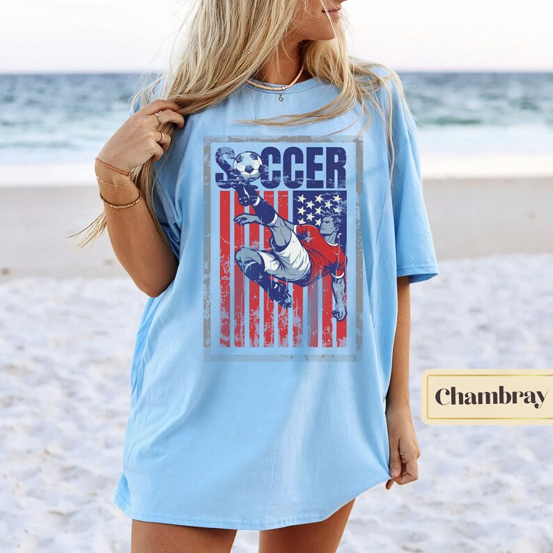 May include: Light blue t-shirt featuring a soccer player kicking a ball, set against a distressed American flag design. The word "SOCCER" is printed above the player. The shirt is a light blue color.
