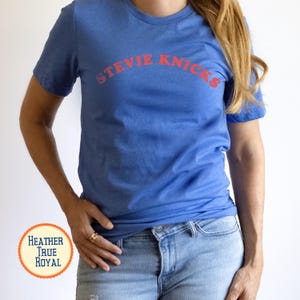 Puede incluir: Una camiseta de manga corta en color azul real jaspeado con el texto STEVIE NICKS impreso en una fuente roja curva en el pecho. La prenda tiene cuello redondo y un corte est&aacute;ndar, combinada con unos pantalones vaqueros de color azul claro.