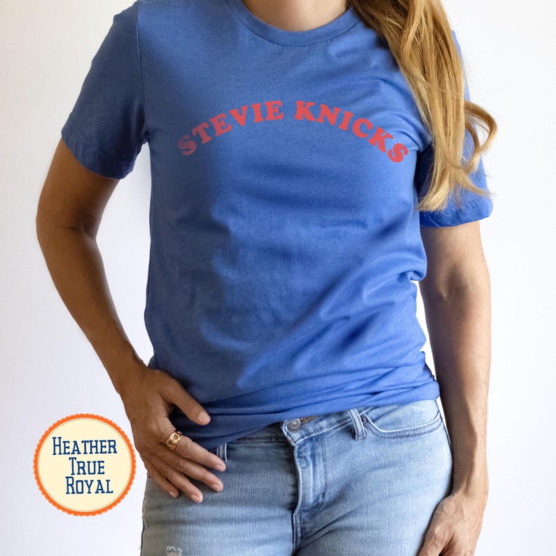 Puede incluir: Una camiseta de manga corta en color azul real jaspeado con el texto STEVIE NICKS impreso en una fuente roja curva en el pecho. La prenda tiene cuello redondo y un corte est&aacute;ndar, combinada con unos pantalones vaqueros de color azul claro.