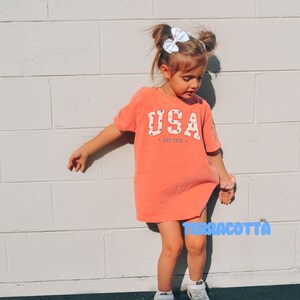 May include: Terracotta-colored t-shirt dress with the word "USA" in white block letters, accented with red and blue stars. The text "EST. 1776" is below the word "USA". The child is wearing white sneakers and a white bow.