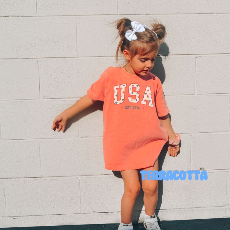 May include: Terracotta-colored t-shirt dress with the word "USA" in white block letters, accented with red and blue stars. The text "EST. 1776" is below the word "USA". The child is wearing white sneakers and a white bow.