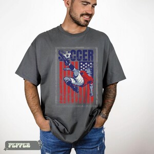 May include: Gray t-shirt with a distressed graphic of a soccer player kicking a ball, set against a red, white, and blue American flag design. The word "SOCCER" is printed above the player.