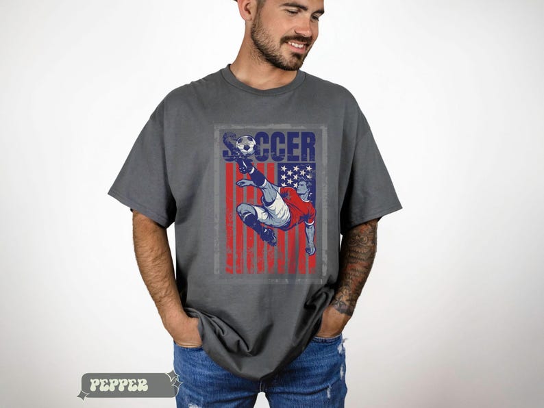 May include: Gray t-shirt with a distressed graphic of a soccer player kicking a ball, set against a red, white, and blue American flag design. The word "SOCCER" is printed above the player.