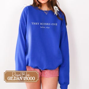 May include: Royal blue Gildan 18000 sweatshirt with the text "THEY MISSED ONE" and "Salem, 1692" in white. The sweatshirt is paired with red and white checkered shorts. The sweatshirt is a long-sleeved, crew neck style.