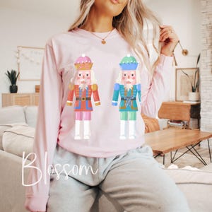 May include: A light pink long-sleeve shirt featuring two cartoon nutcracker figures. One nutcracker is red and gold, the other is blue and green. The word "Blossom" is written in cursive at the bottom of the shirt.