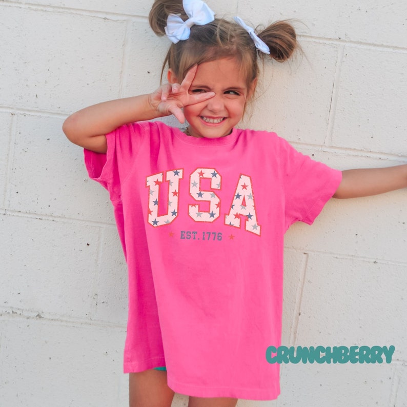 May include: A bright pink t-shirt with the word "USA" in a collegiate style, featuring red, white, and blue stars. The text "EST. 1776" is below the word "USA". The shirt is worn by a young person.