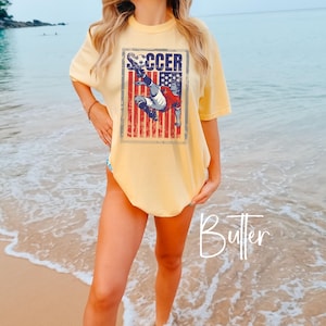 May include: Butter-colored t-shirt with a soccer graphic. The graphic features a soccer player kicking a ball in front of a distressed American flag design. The word "SOCCER" is printed above the player. The shirt has short sleeves and a relaxed fit.