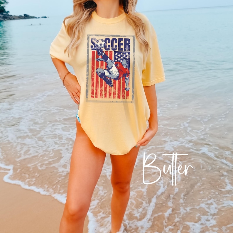 May include: Butter-colored t-shirt with a soccer graphic. The graphic features a soccer player kicking a ball in front of a distressed American flag design. The word "SOCCER" is printed above the player. The shirt has short sleeves and a relaxed fit.
