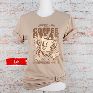 May include: Tan t-shirt with a retro cartoon coffee cup graphic. The text reads "Sunshine Coffee Shop Est. 1958 Coffee Break More Espresso Less Depresso".