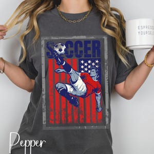 May include: Dark gray t-shirt featuring a graphic of a soccer player kicking a ball, set against a distressed American flag design. The word "SOCCER" is printed above the player. The shirt has a vintage, worn look.