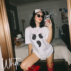May include: A white short-sleeved t-shirt featuring a black graphic of a ghost face with long eyelashes. The outfit includes a black and white baseball cap, dark sunglasses, and red cowboy boots. The shirt has the word White printed in script.