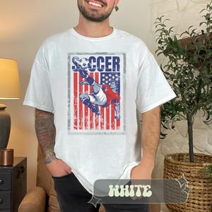 May include: White t-shirt featuring a distressed graphic of a soccer player kicking a ball, set against a backdrop of the American flag. The word "SOCCER" is printed above the image. The word "WHITE" is printed at the bottom of the shirt.