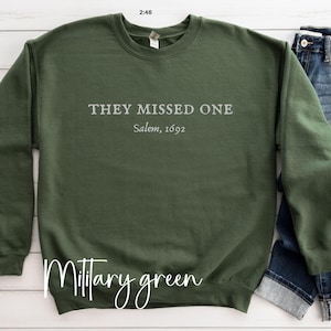 May include: A military green sweatshirt with the text "They Missed One Salem, 1692" printed on the front.