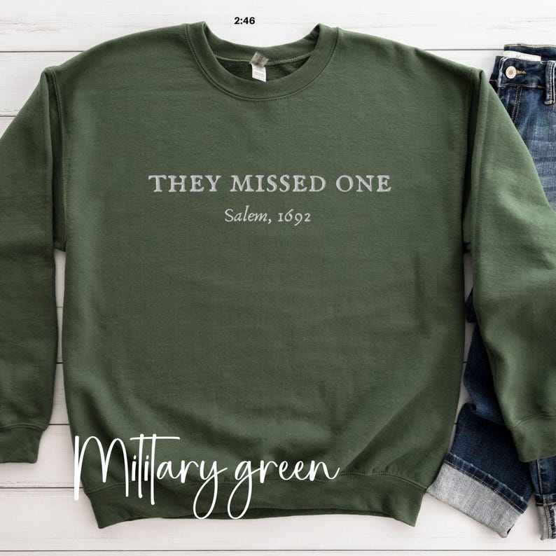May include: A military green sweatshirt with the text "They Missed One Salem, 1692" printed on the front.