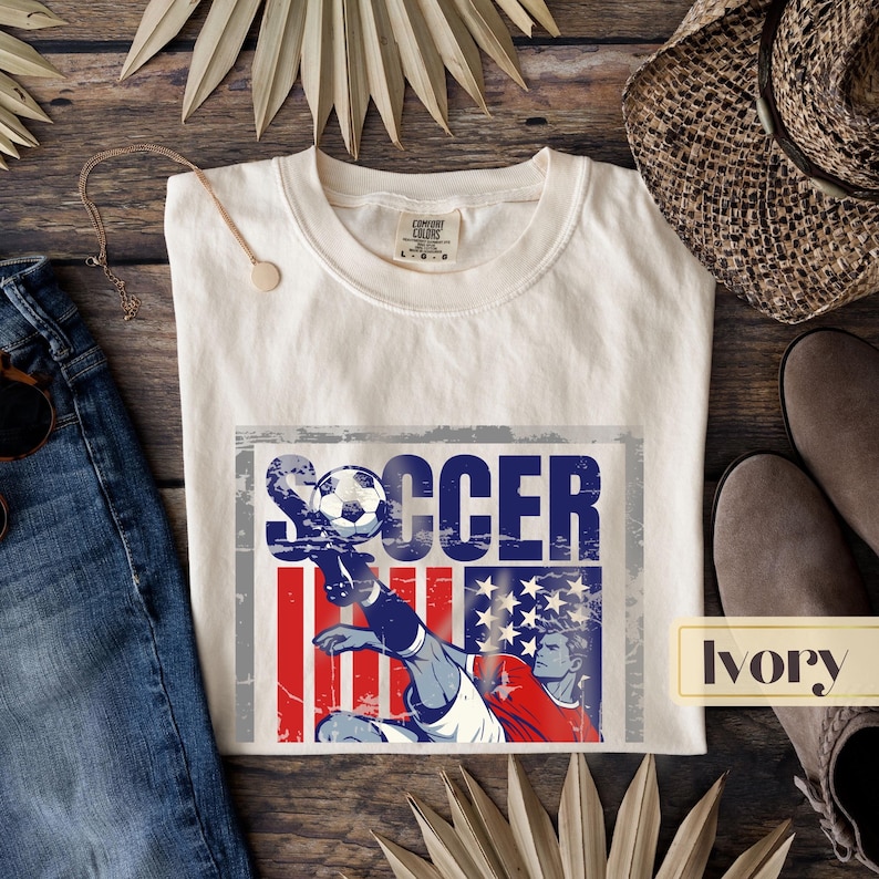 May include: Ivory-colored t-shirt with a distressed graphic featuring the word "SOCCER" and a soccer player kicking a ball. The graphic incorporates red, white, and blue elements. The shirt is displayed on a wooden surface with other accessories.