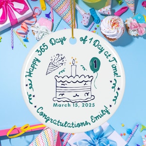 May include: A white ceramic ornament with a birthday cake illustration, a green number one balloon, and the text "Happy 365 Days of 1 Day at a Time!" and "Congratulations, Emily!" The date "March 15, 2025" is also included.