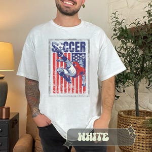 May include: White t-shirt with a distressed graphic featuring a soccer player kicking a ball, set against a backdrop of the American flag. The word "SOCCER" is printed above the image. The shirt is a casual, unisex style.